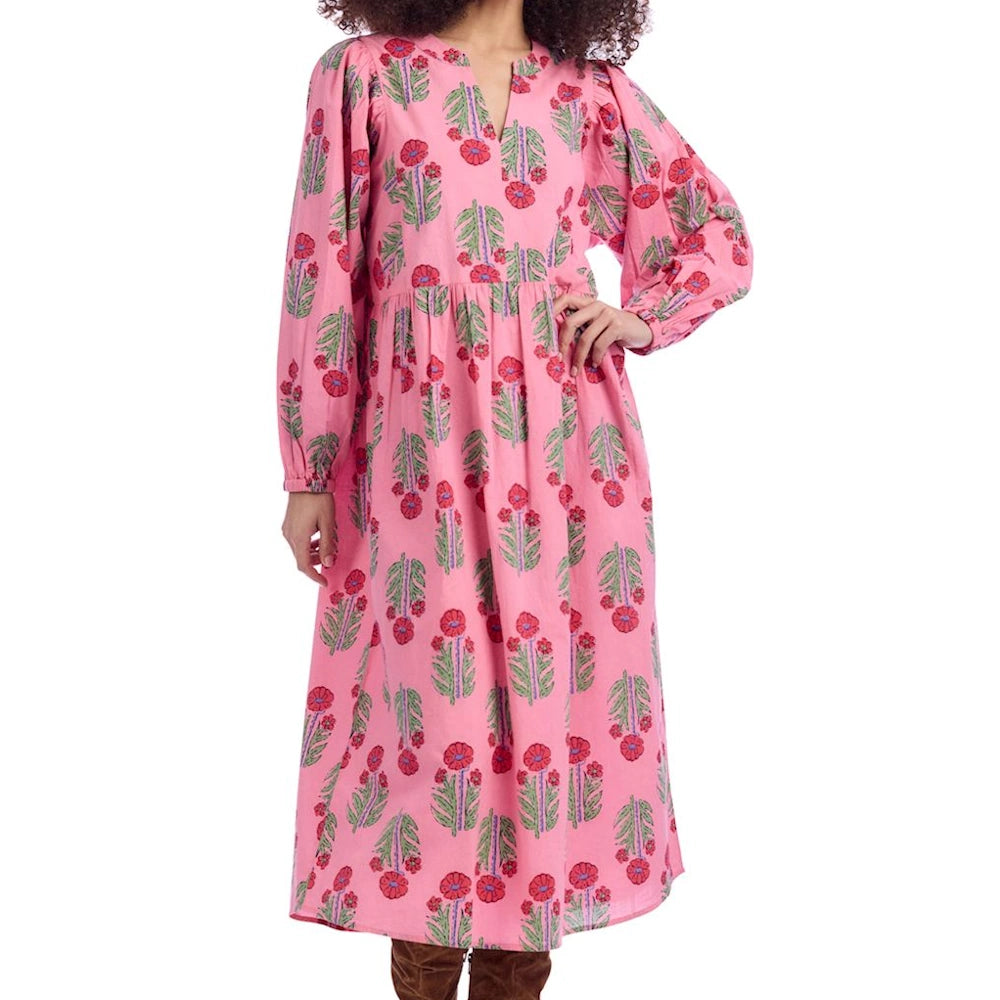 A woman wears a pink long-sleeve midi dress with a red floral block print, V-neckline, and tiered skirt against a white background.