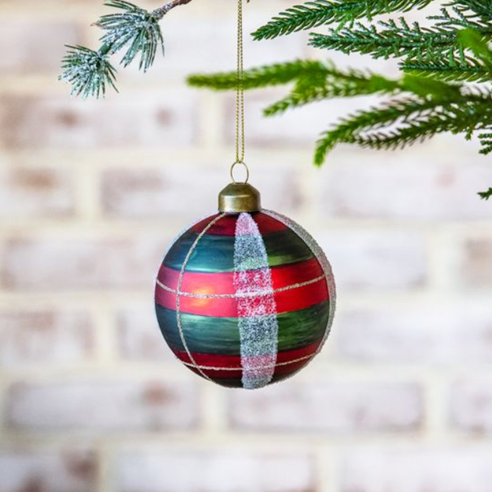 Red and green plaid glass ball ornament with frosted glitter stripes hanging on a pine branch.
