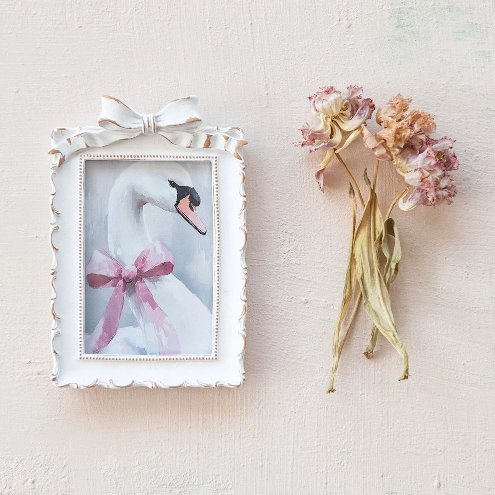 White ornate picture frame with a swan painting and pink bow, next to dried pink flowers on a textured cream wall.