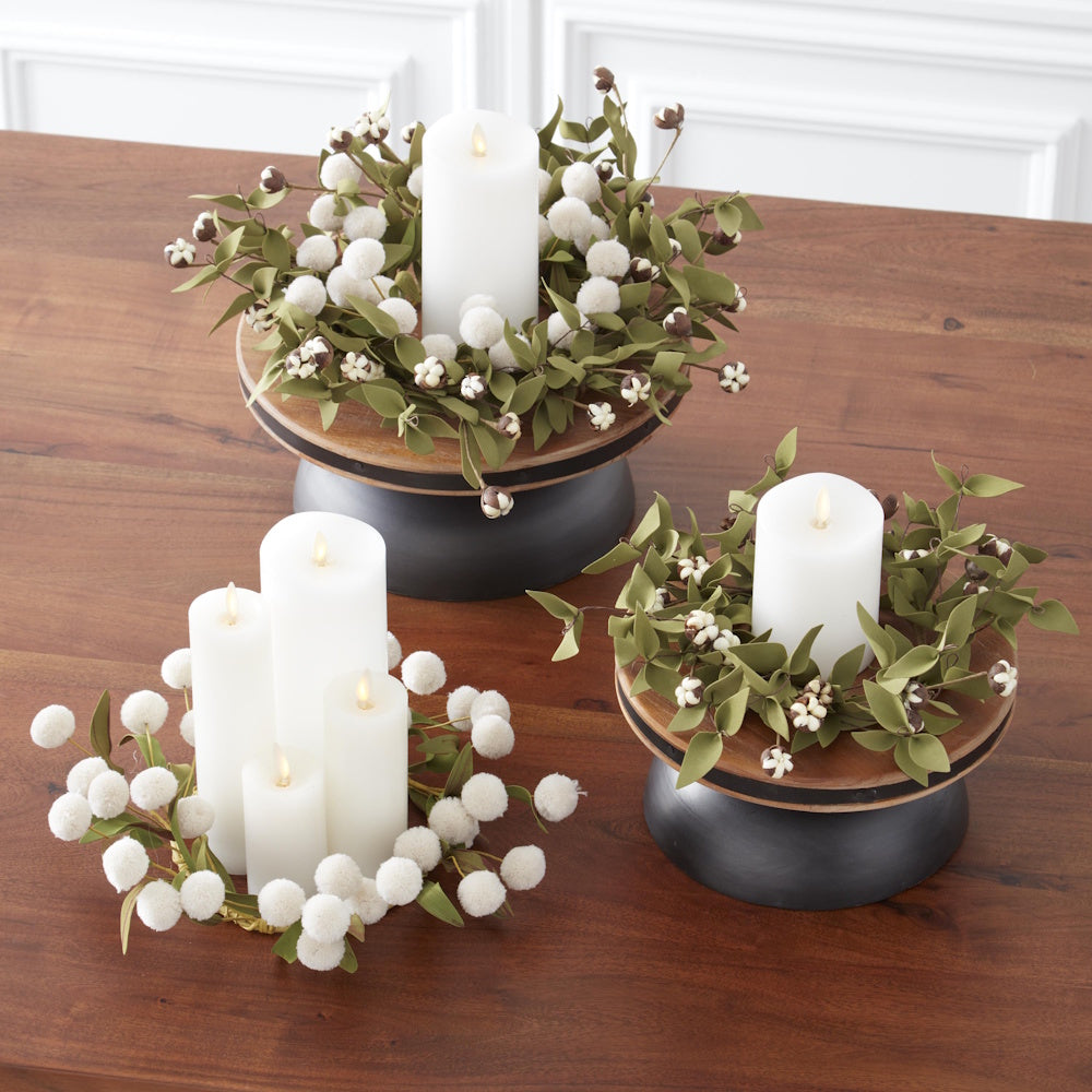 Three white candle arrangements with green foliage and white pom-poms on a wooden table.