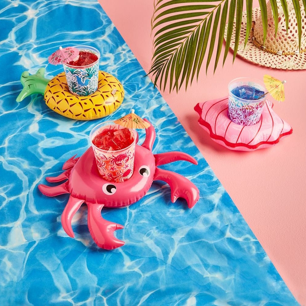 Inflatable crab, pineapple, and seashell drink floats holding iced cocktails on blue and pink surfaces.