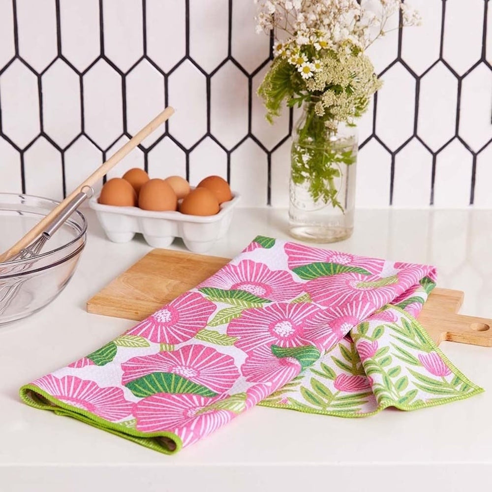 Pink poppy print tea towel on a wooden board next to a bowl, eggs, and a floral vase in a kitchen.