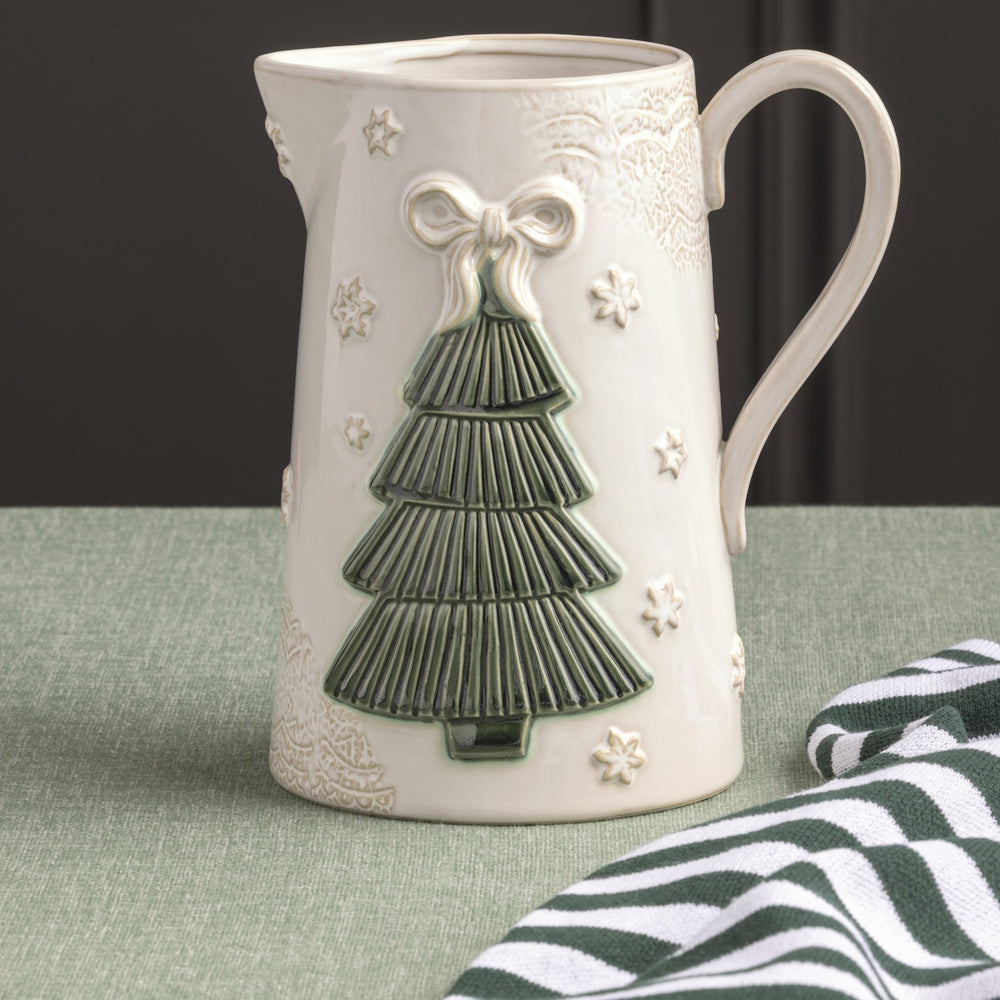 Front view of a cream ceramic Christmas tree pitcher styled on a table with a green tablecloth and striped towel.