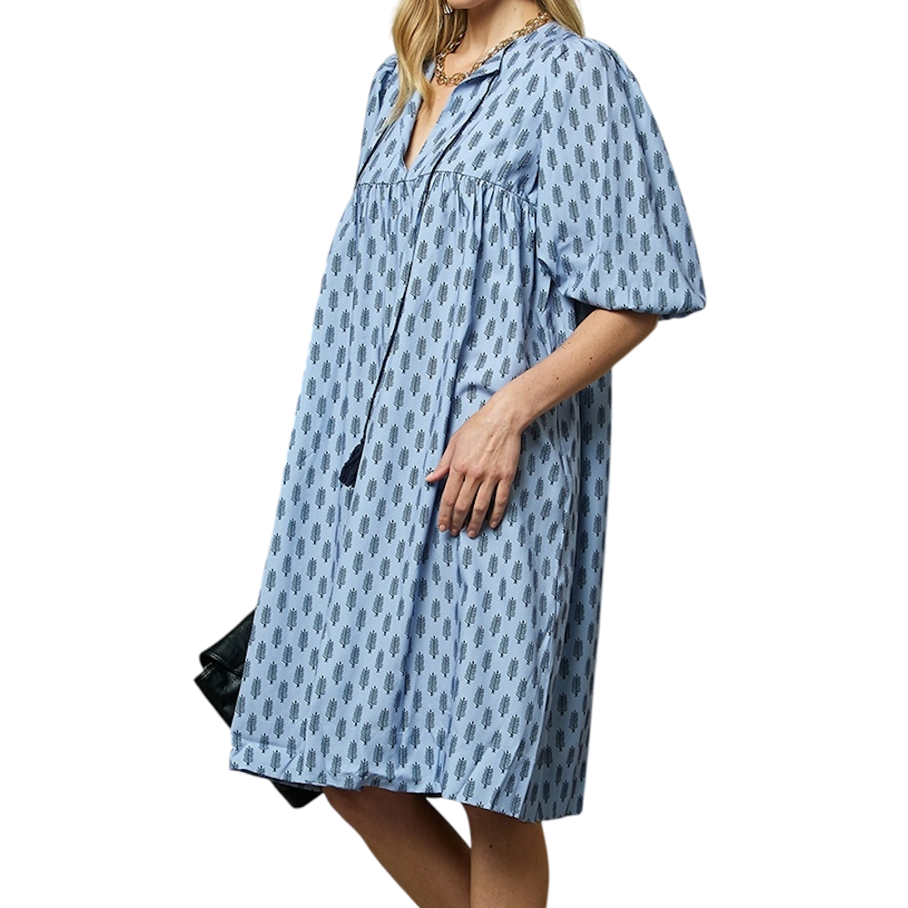 A woman wearing a light blue, knee-length dress with puff sleeves and a repeating dark block print pattern.