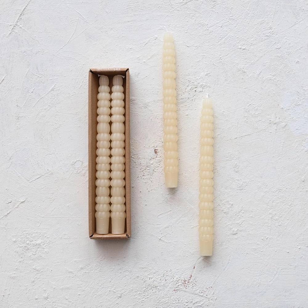 Four ivory taper candles with a textured, ribbed design, shown both inside a cardboard box and loosely alongside.