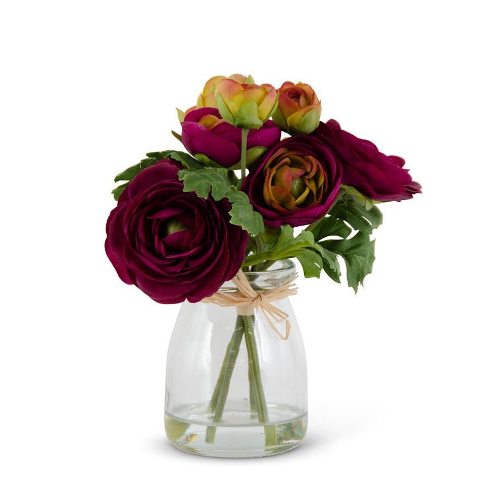 A small bouquet of deep magenta and yellow ranunculus flowers in a clear glass jar vase on a white background.