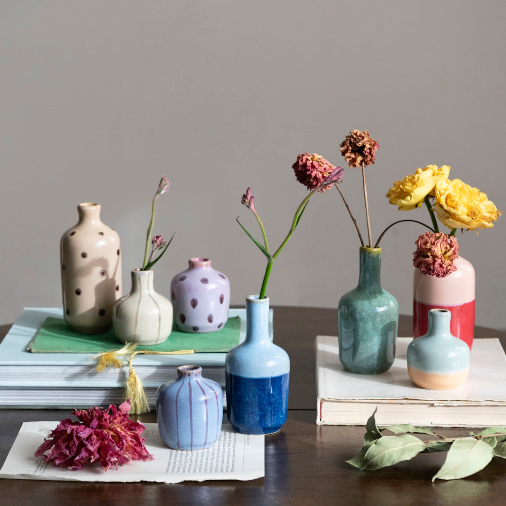 Assorted small, colorful stoneware vases with flowers arranged on books and a dark wooden table.