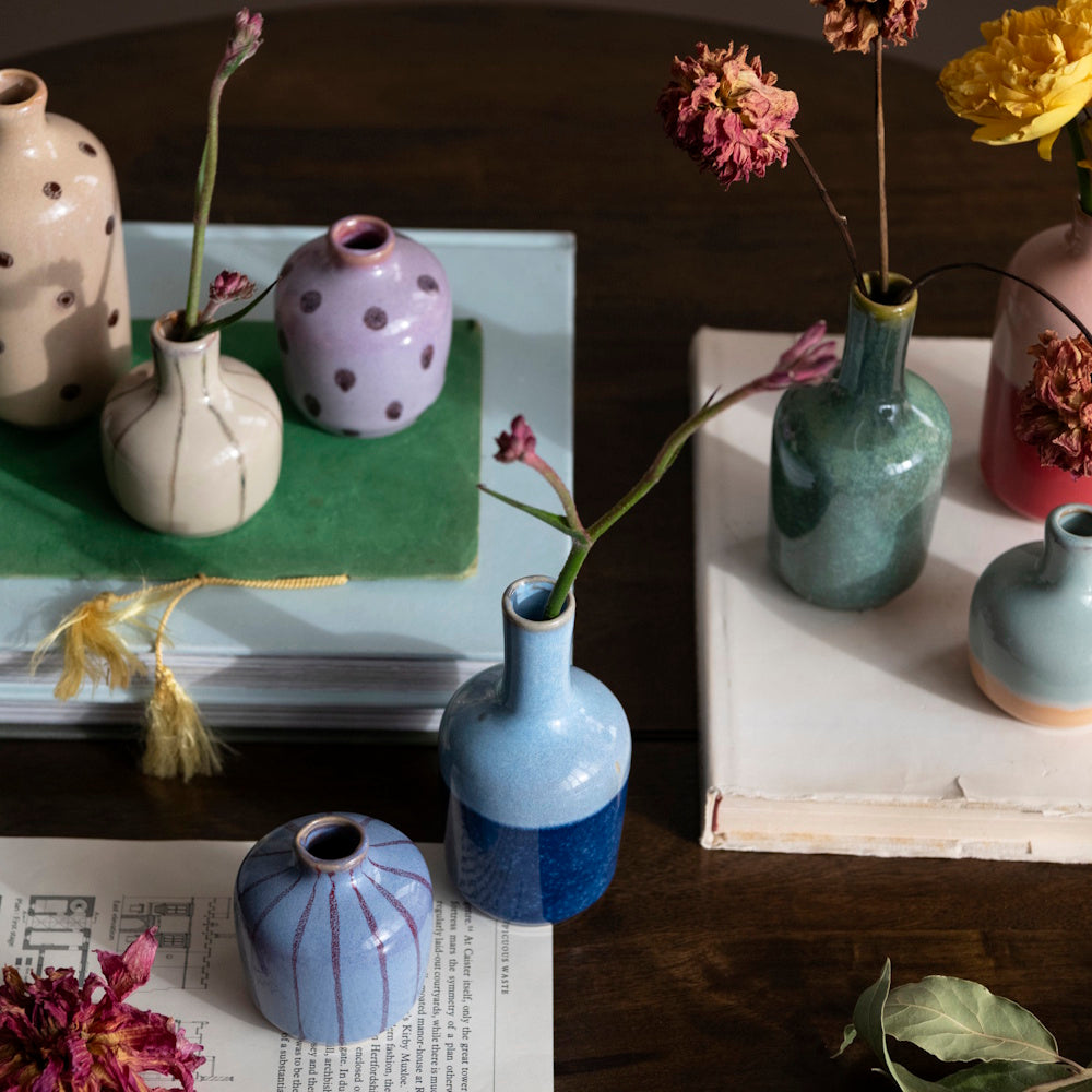 An assortment of small, colorful hand-painted stoneware bud vases with dried florals on a wooden table.