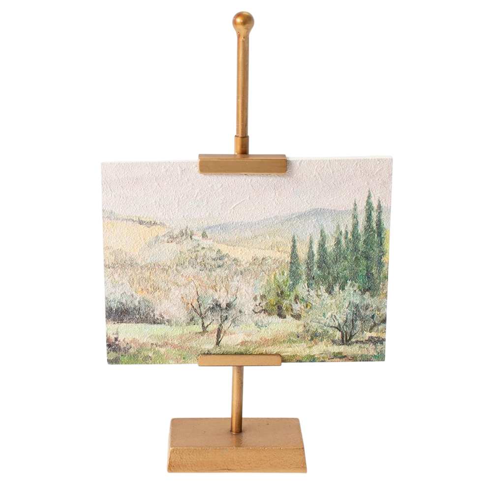 Mini landscape painting of a countryside valley displayed on a gold metal easel stand against white background.