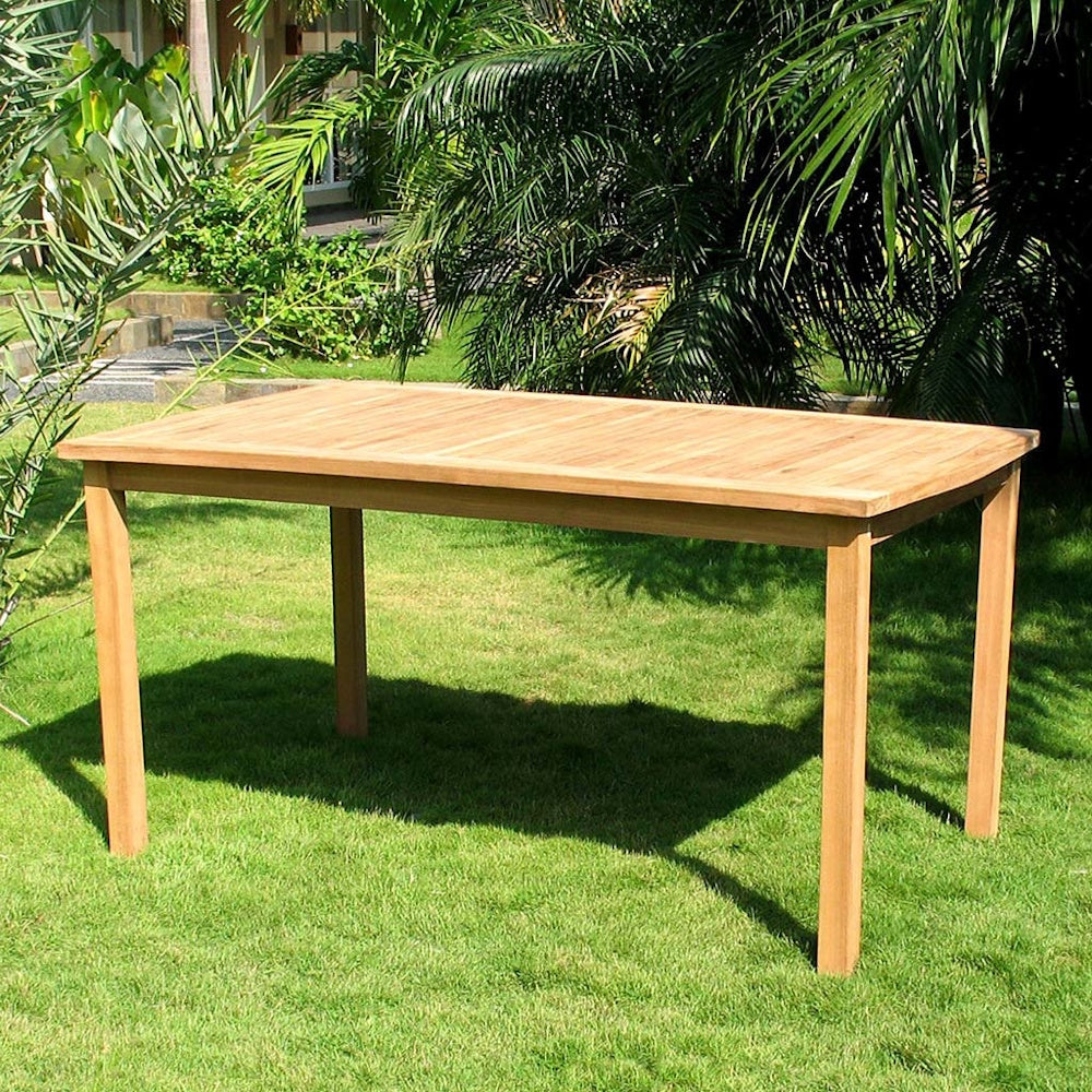 Rectangular slatted teak wood outdoor dining table set on a grassy lawn surrounded by tropical foliage.