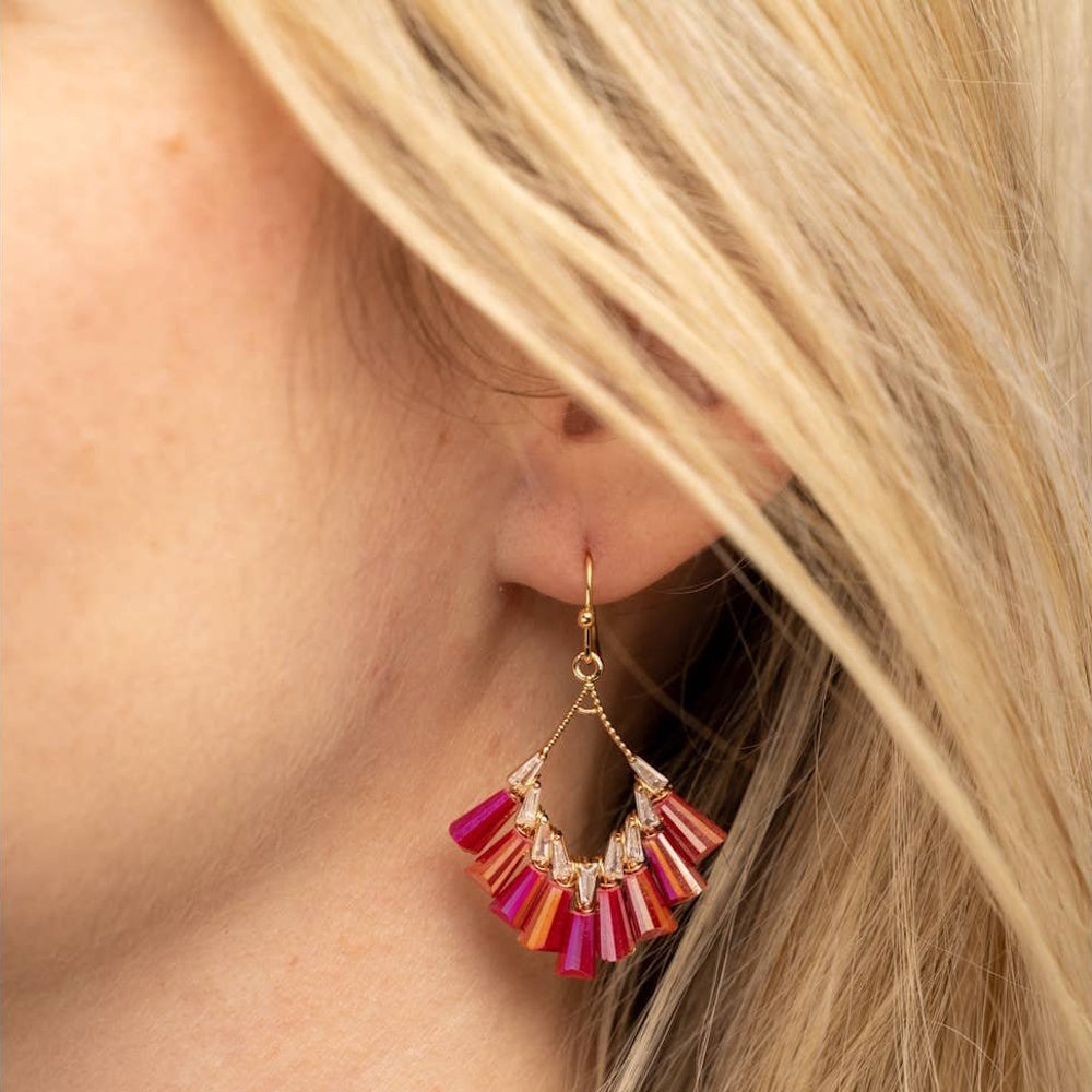 A person with blonde hair wearing a gold dangle earring featuring a fan of pink and reddish-orange crystals.