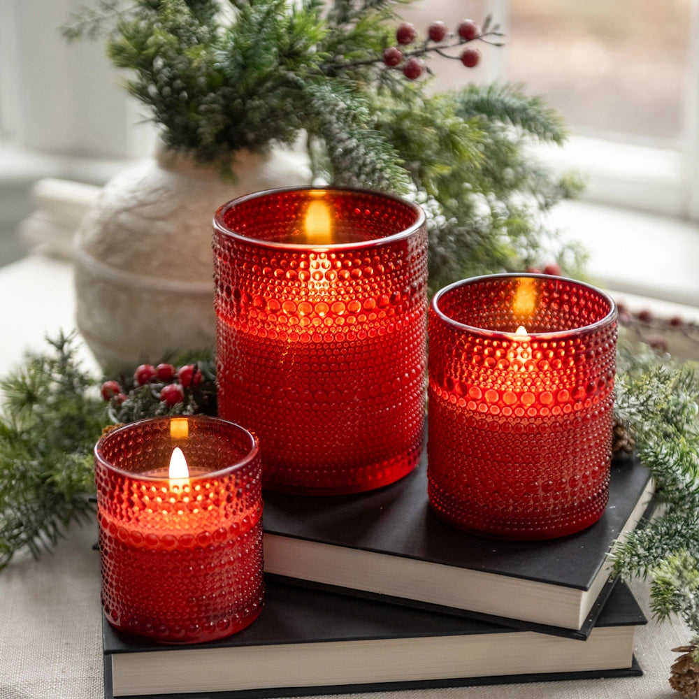 Three lit red beaded glass candles in varying sizes set on stacked books with holiday greenery.