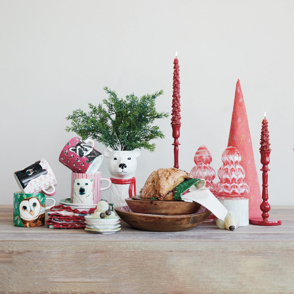 A festive holiday tablescape featuring animal-themed mugs, a polar bear vase, wooden bowls holding pastries, and red taper candle holders.