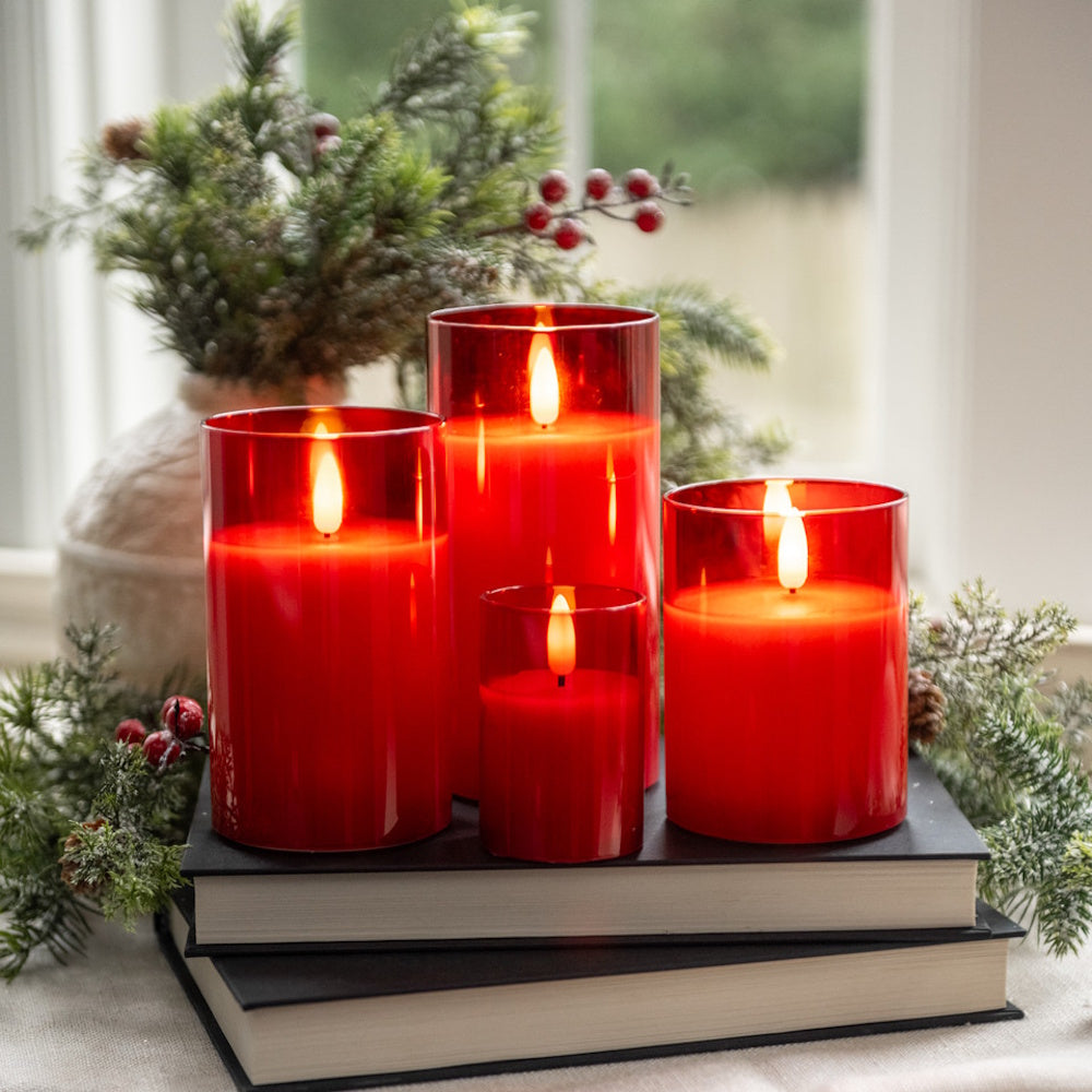 Four glowing red glass LED candles stacked on books near evergreen pine branches in front of a window.