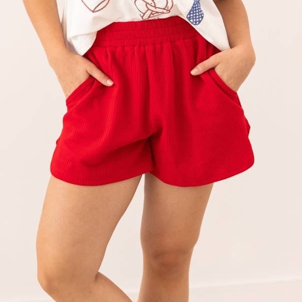 A person poses with hands in the pockets of bright red, high-waisted ribbed knit shorts with an elastic band.
