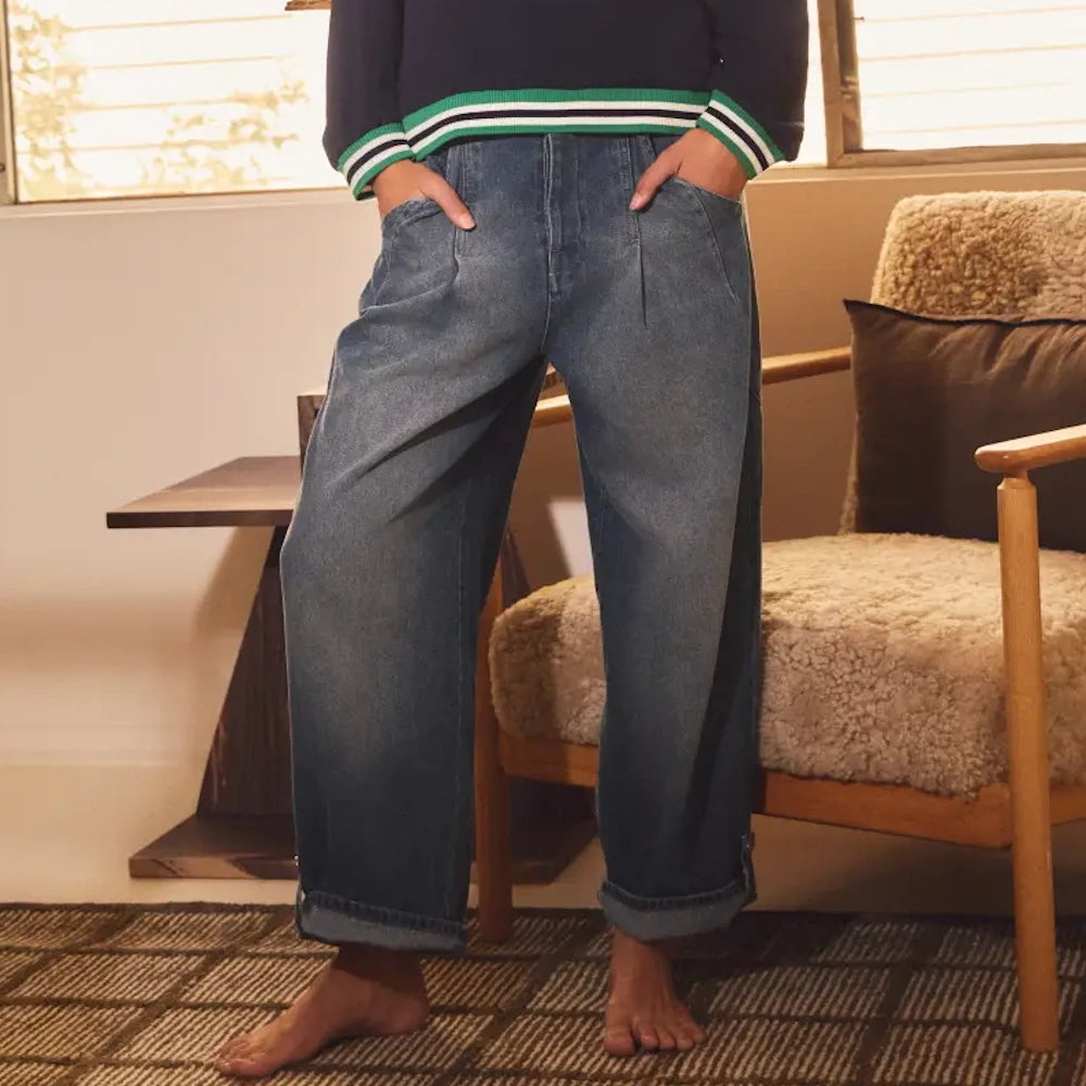Person wearing relaxed-fit, wide-leg denim jeans with cuffed hems and front pleats in a cozy indoor setting.