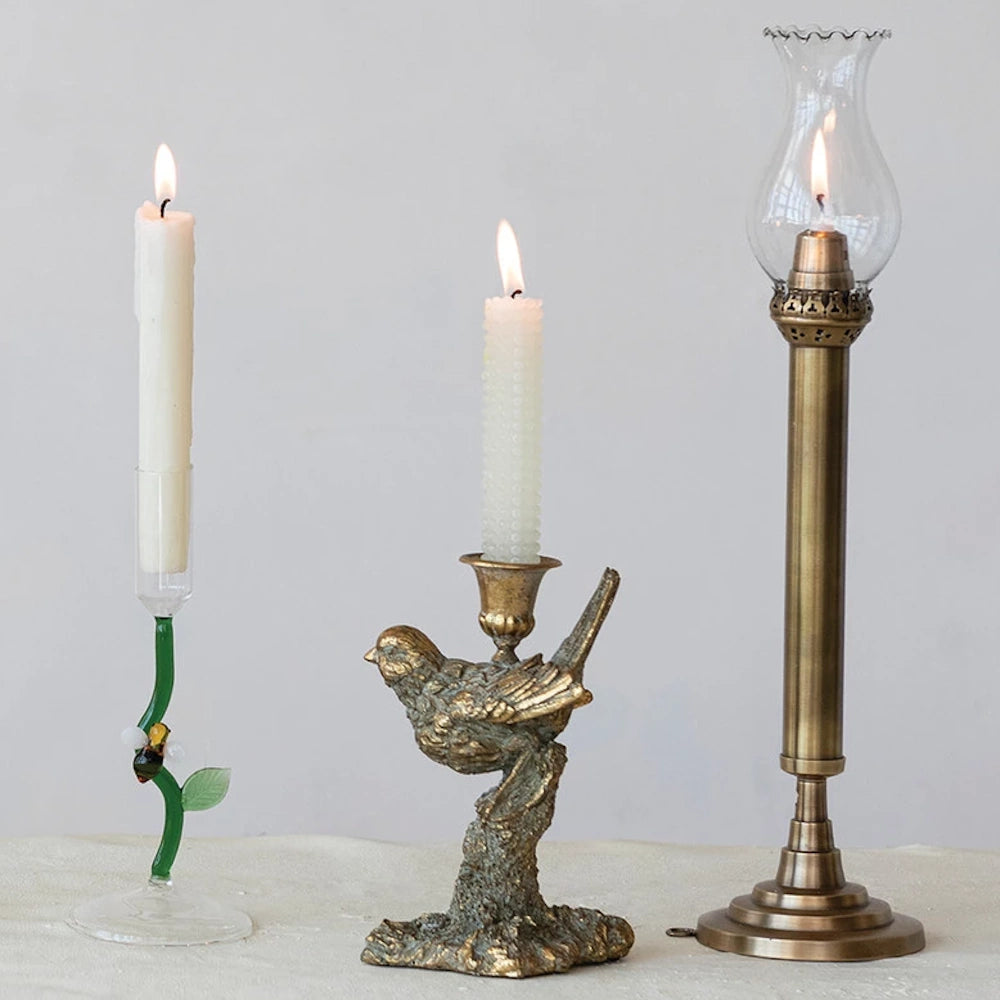 Three unique vintage-style candle holders featuring a glass vine, a bronze bird on a branch, and a tall brass hurricane lamp.