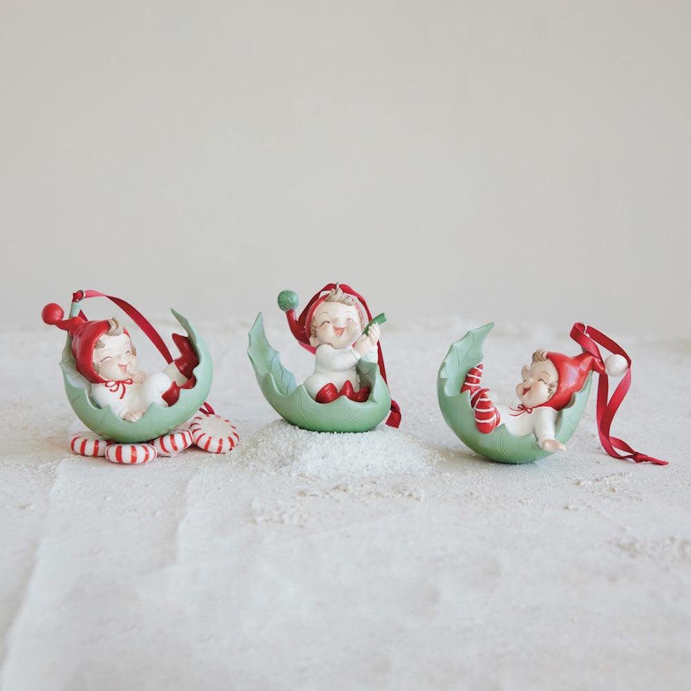 Three resin elf ornaments resting in green leaf-shaped cradles with red ribbons.