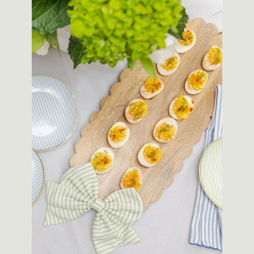 Deviled eggs with dill and paprika on a scalloped wooden serving board with a green striped bow.