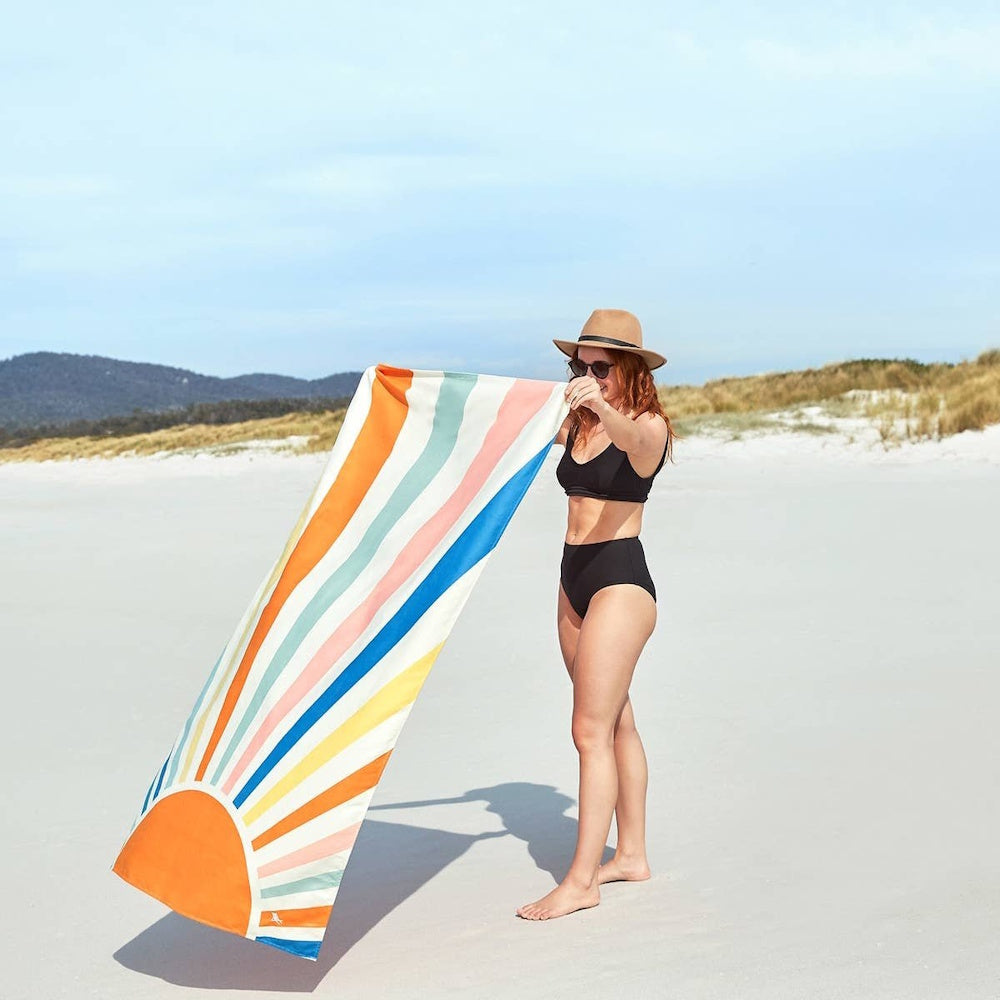 Woman in a black bikini and hat holds up a colorful, striped rising sun beach towel on a white sand beach.