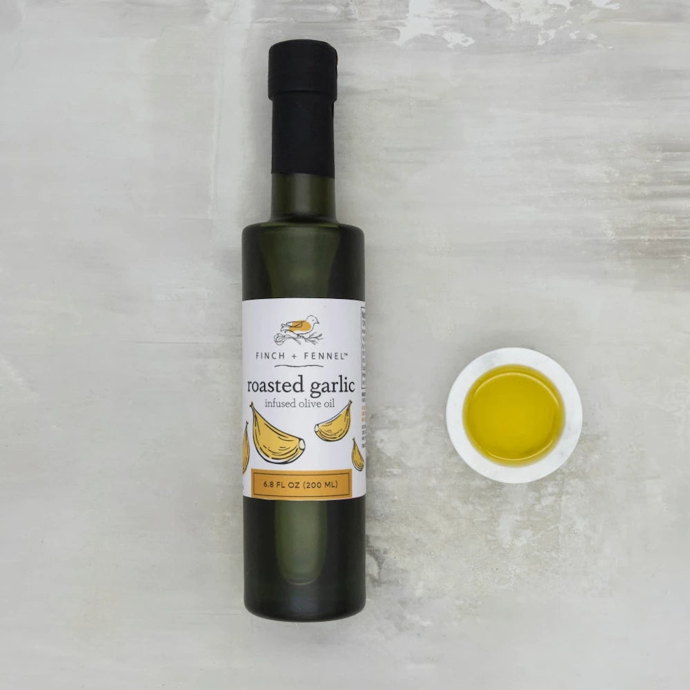 Finch + Fennel roasted garlic infused olive oil bottle next to a small bowl of golden oil on a gray background.