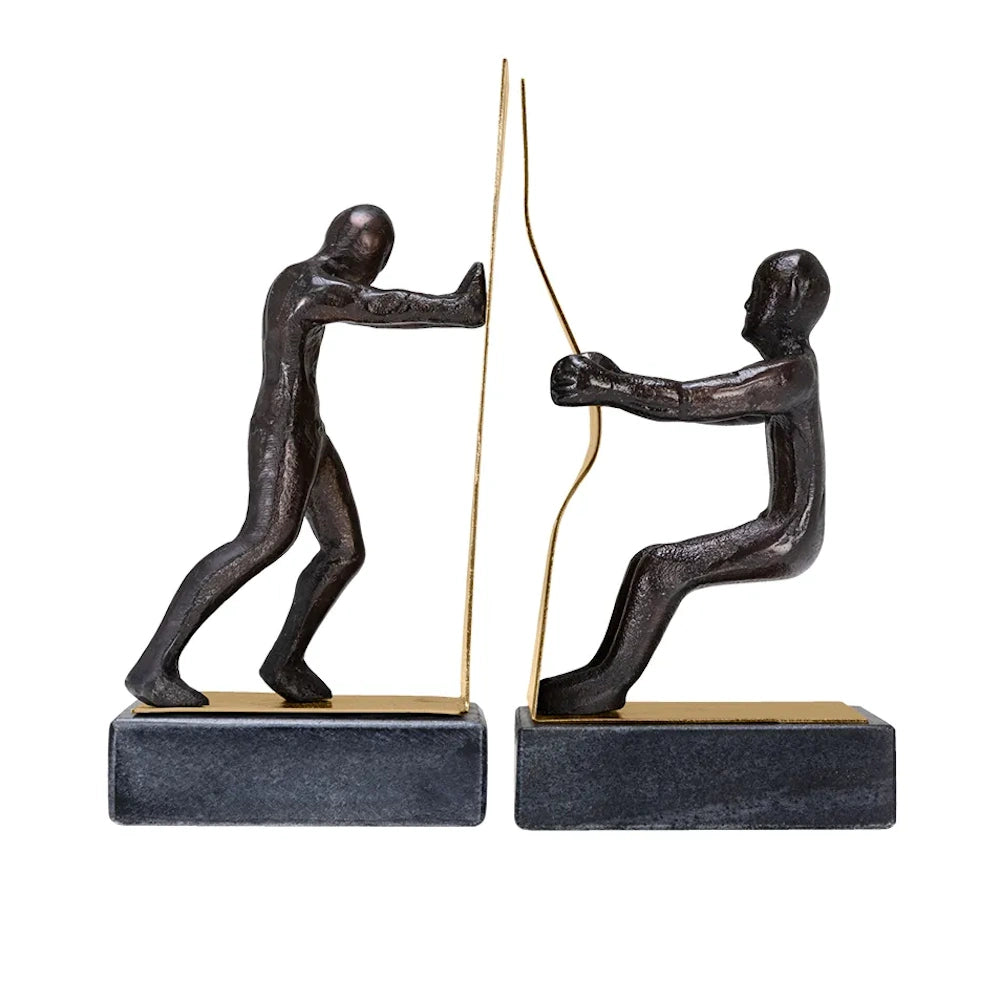 Two bronze figure bookends on dark bases, one pushing a gold plate and the other pulling a wavy gold ribbon.