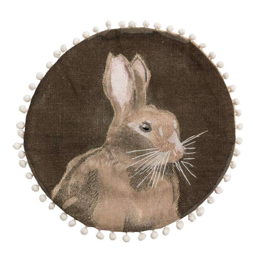 Front view of a round dark brown cushion featuring an embroidered rabbit with a cream pom-pom trim border.
