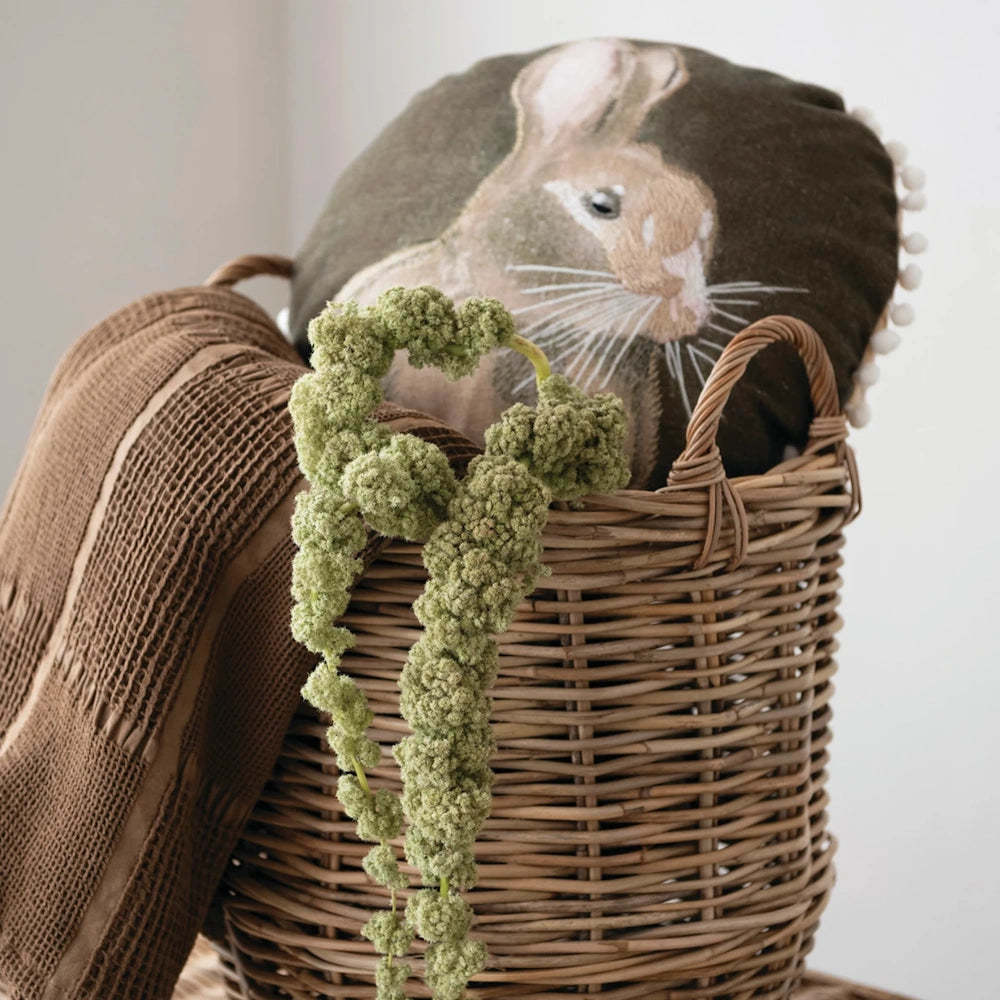 Round rabbit pillow tucked into a woven basket with a textured brown blanket and green greenery.