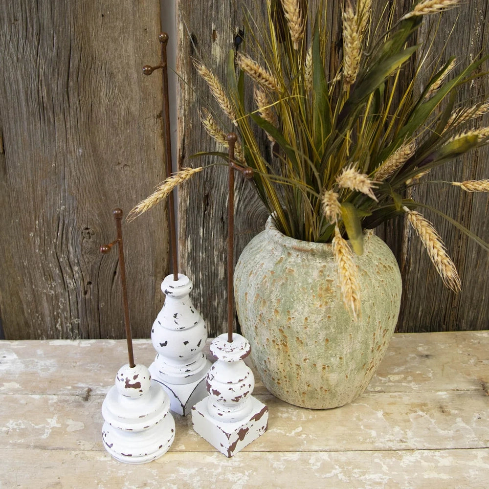 Front view of three rust metal charm display stakes set in distressed white bases on a rustic table.