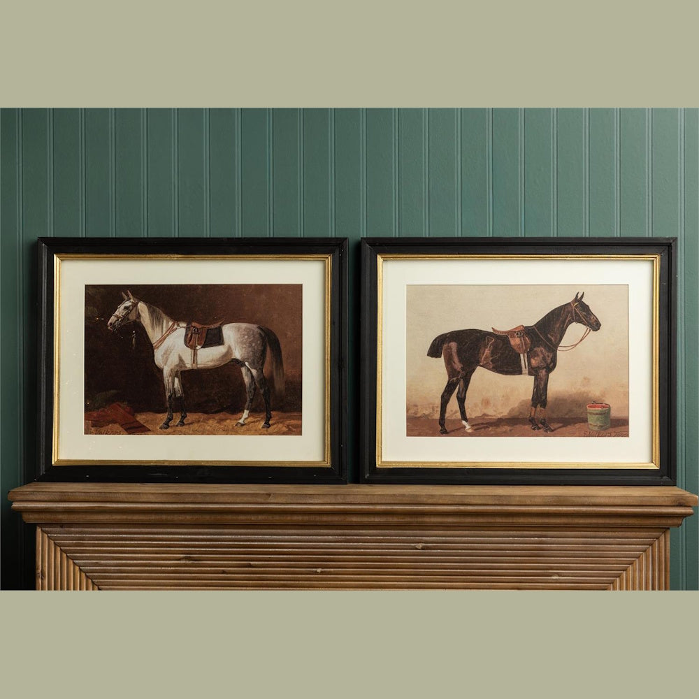 Two framed paintings of saddled horses, one white and one dark brown, resting on a wooden mantelpiece.