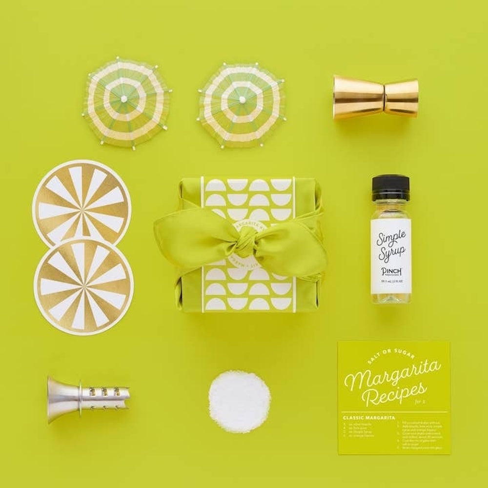 Margarita cocktail kit with a lime-green wrapped gift, syrup, recipes, and cocktail accessories on a green background.