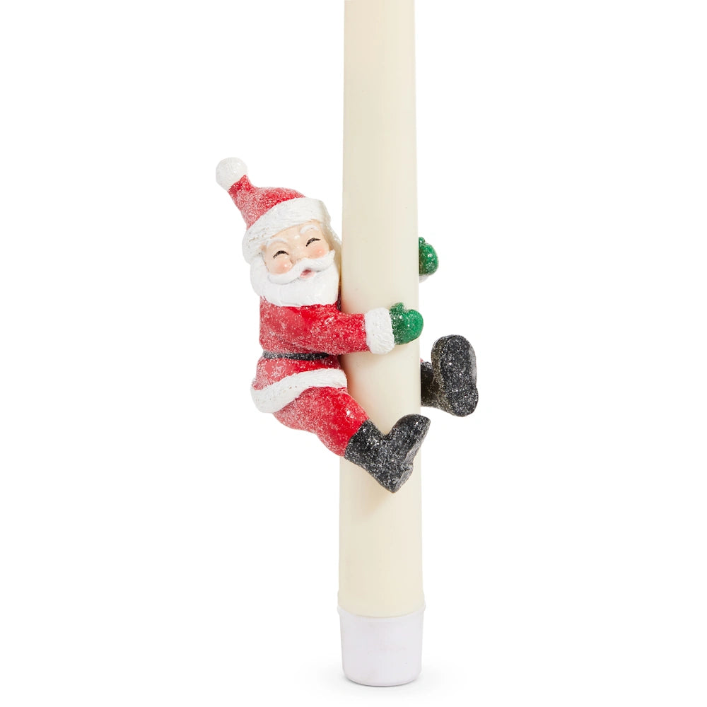 A Santa Claus figurine wearing a red suit and green mittens hugging a tall ivory candle.