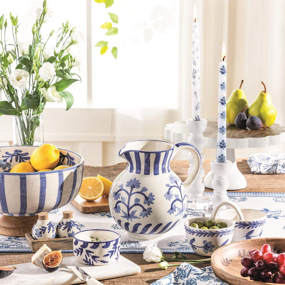 Blue and white floral ceramic pitcher, bowls, and taper candles on a rustic dining table with fruit and flowers.