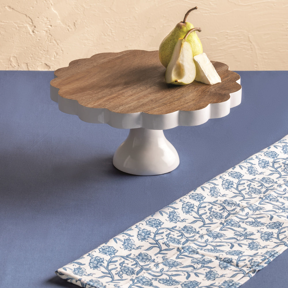 Scalloped wood and white ceramic pedestal cake stand with pears and brie on a blue table with a floral napkin.