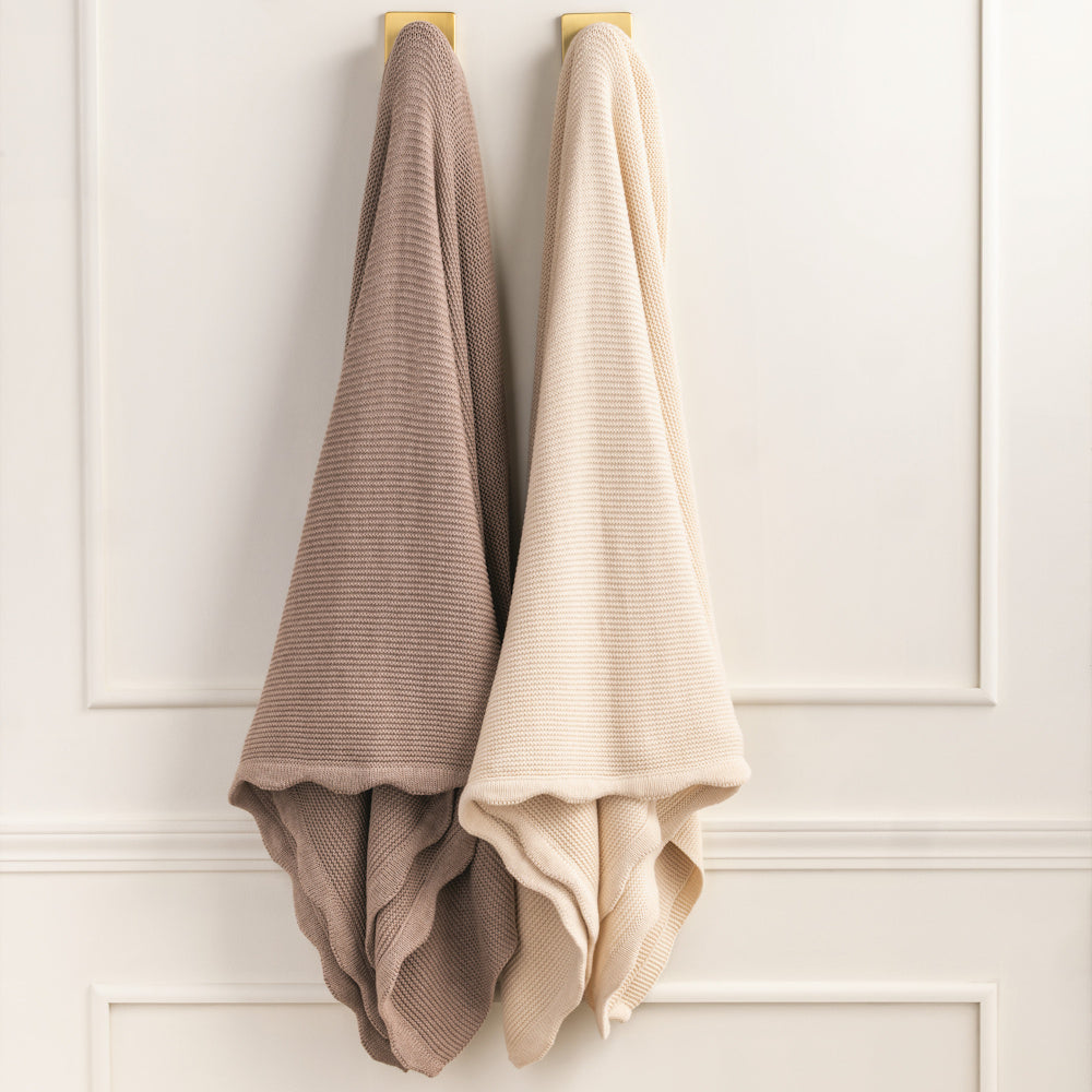 Two textured knit scalloped throw blankets in mauve and cream hanging on gold hooks against a white paneled wall.