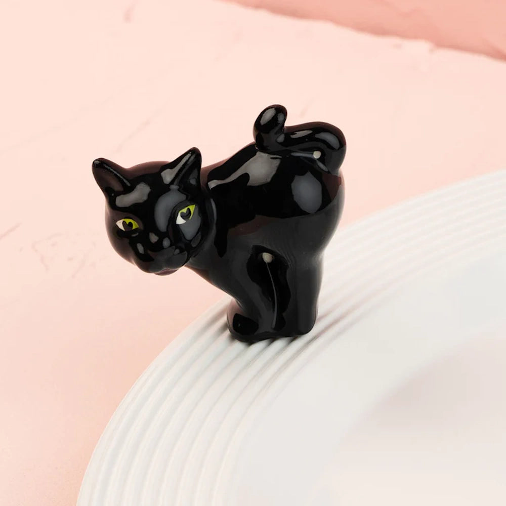 A small, glossy black cat figurine with green eyes perches on the ridged rim of a white ceramic plate.