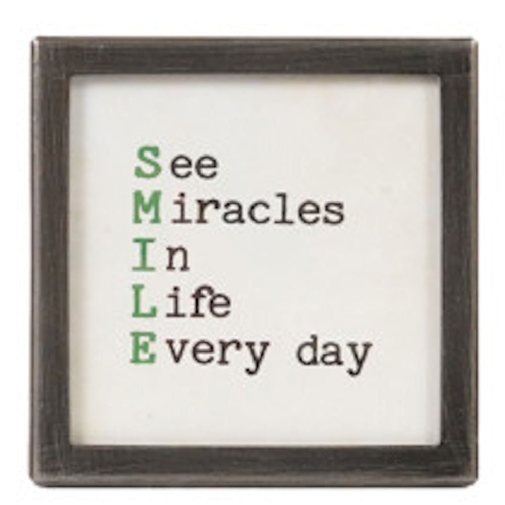 A square silver-framed sign with the text "See Miracles In Life Every day" where the first letters spell "SMILE" in green.