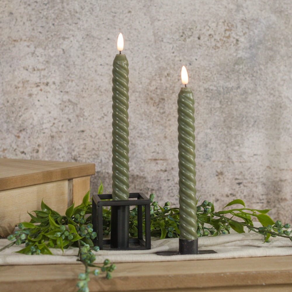 Two glowing dark green spiral twisted taper candles in black holders set among leafy greenery and berries.