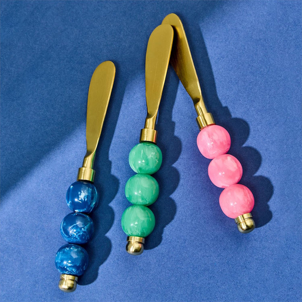 Three gold butter spreaders with blue, green, and pink beaded handles on a blue background.