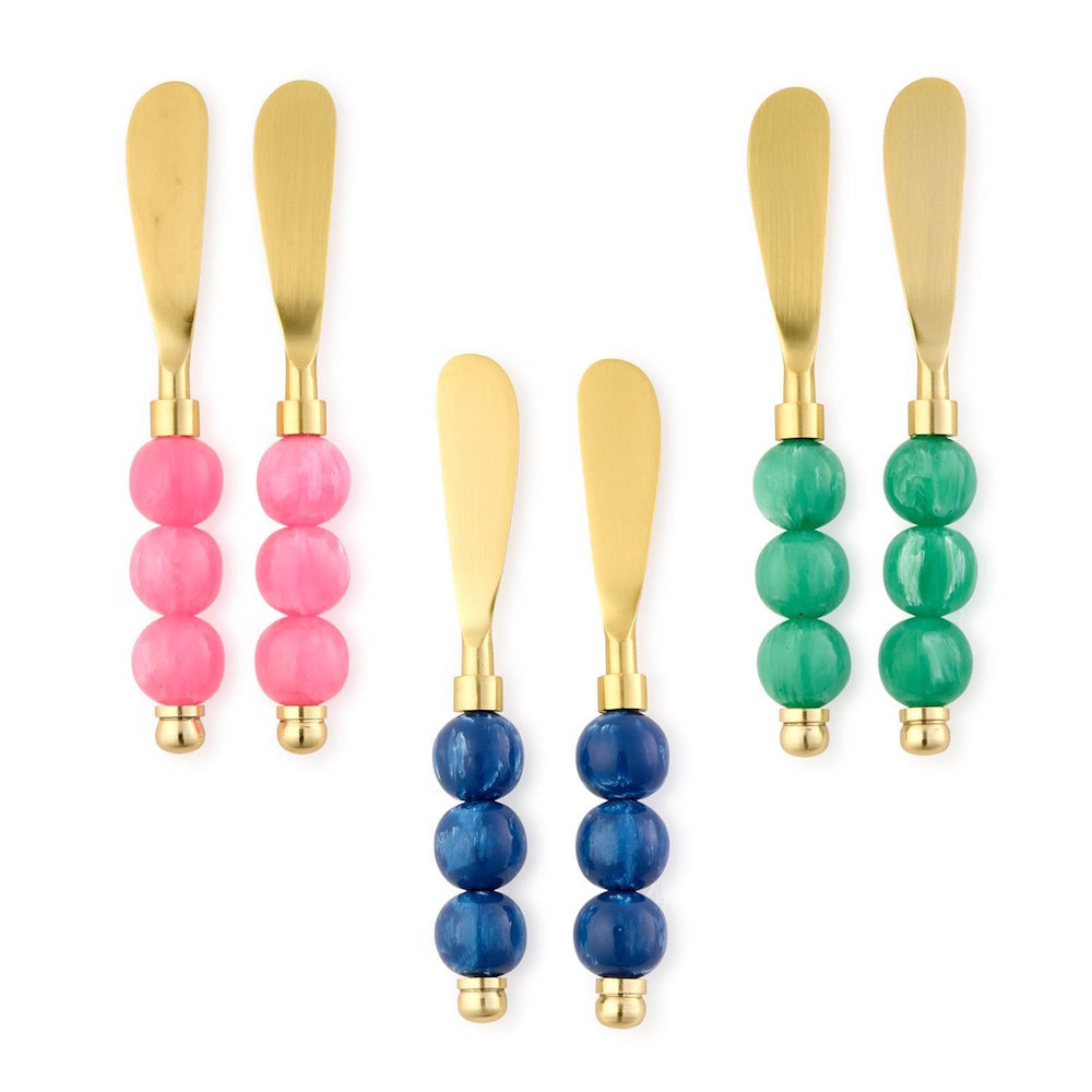 Three pairs of gold cheese spreaders with colorful beaded handles in pink, blue, and green on a white background.