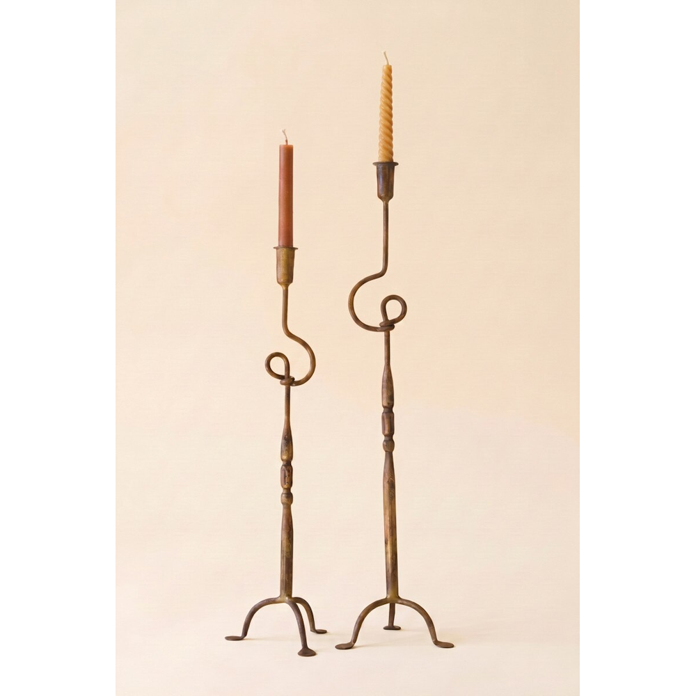 Two slender antique brass taper candleholders with twisted loop details standing on a beige background.