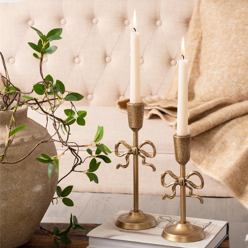 Two gold taper candlestick holders featuring decorative metal bow accents holding lit white candles.