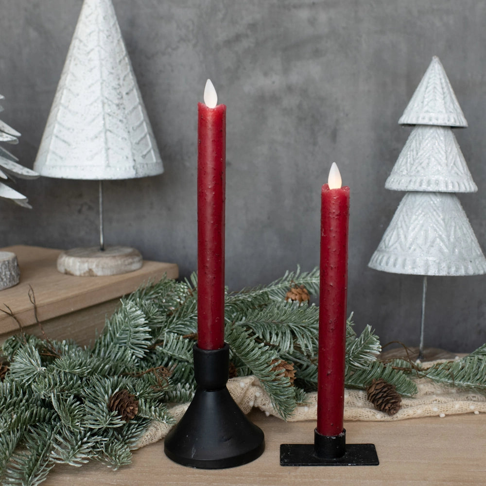 Two red taper candles in black holders surrounded by evergreen branches and white tree decor.