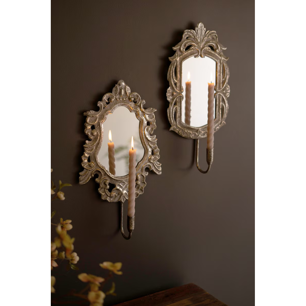 Angled view of two vintage-style gold mirror wall sconces with glowing candles on a dark wall.