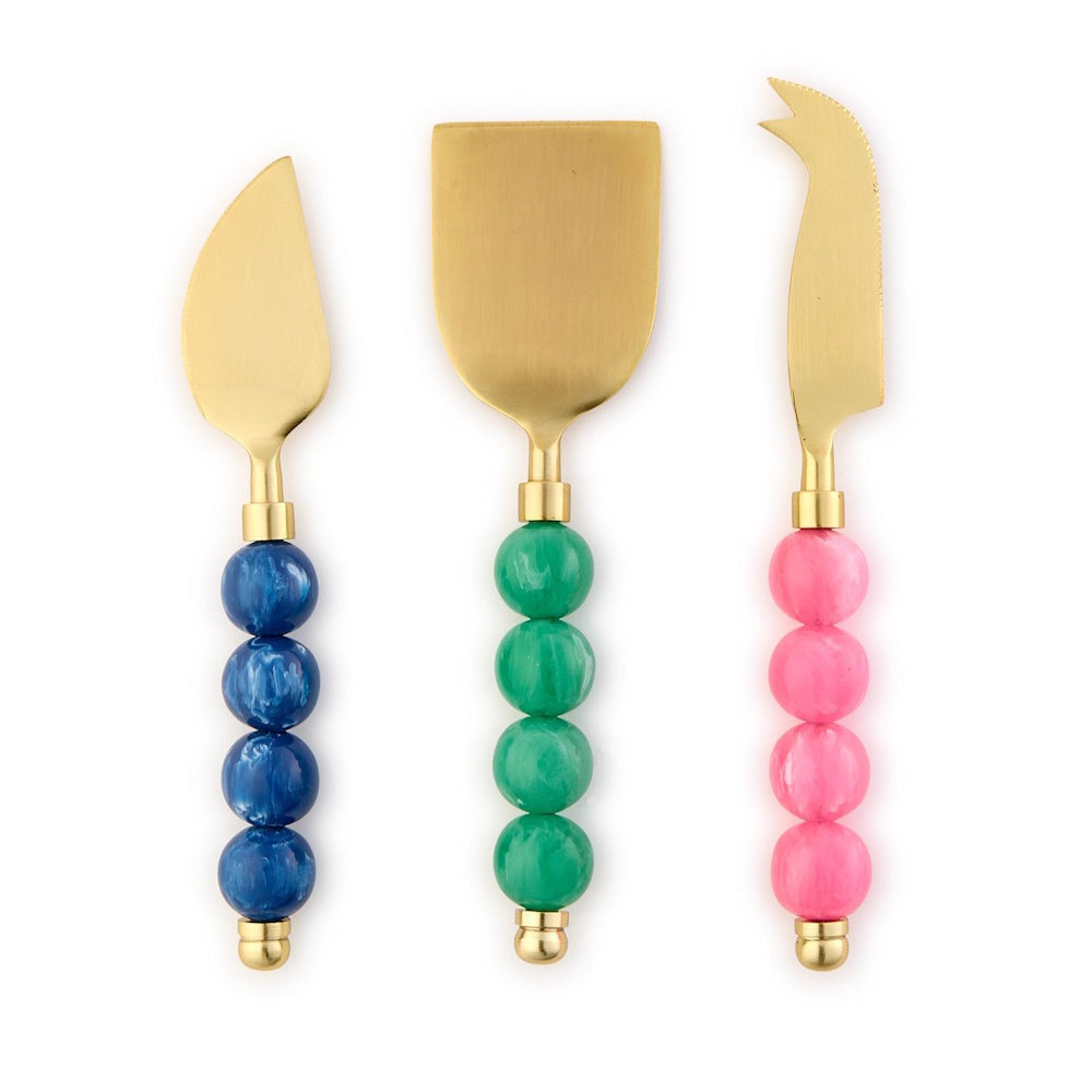 Set of three gold cheese knives with colorful beaded handles in blue, green, and pink on a white background.