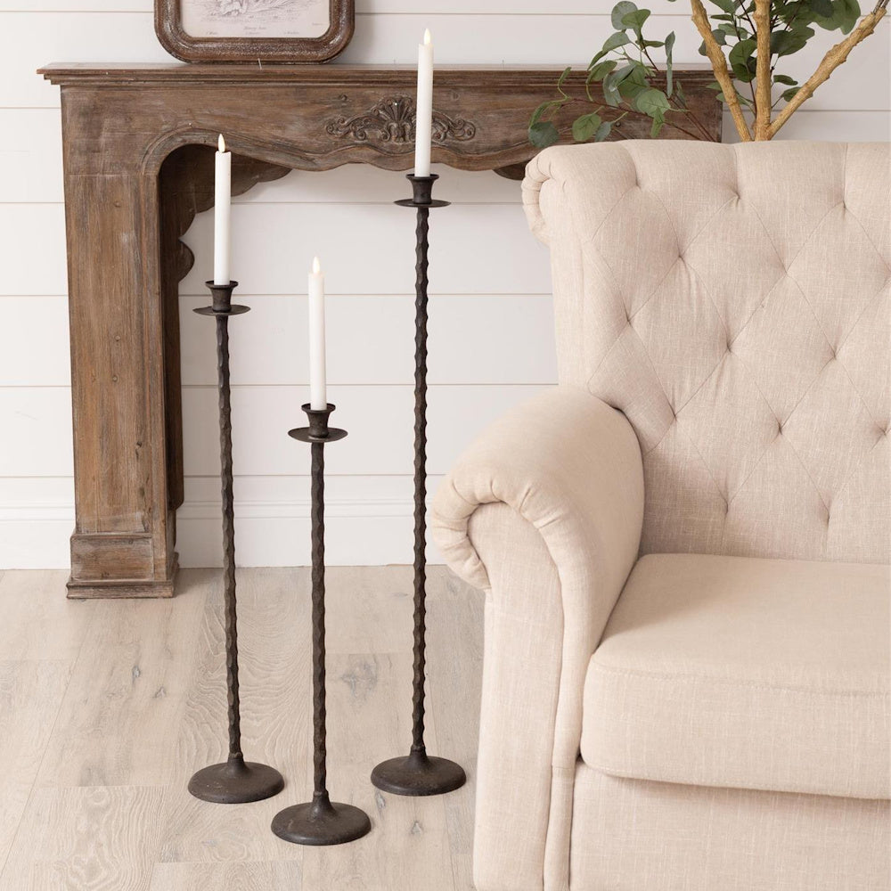 Set of 3 rustic wrought iron floor taper candle holders in a farmhouse living room next to a beige tufted chair.
