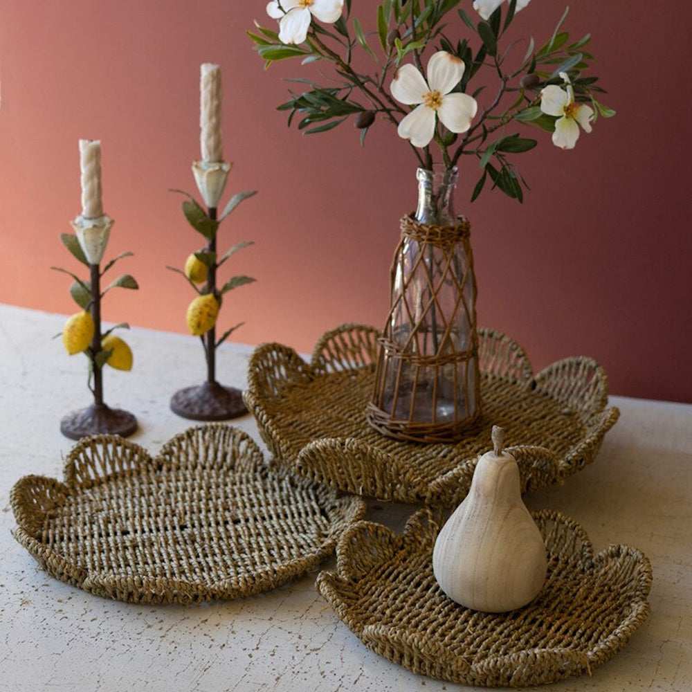 Set of three flower-shaped woven seagrass trays styled with a flower vase, candles, and a wooden pear.