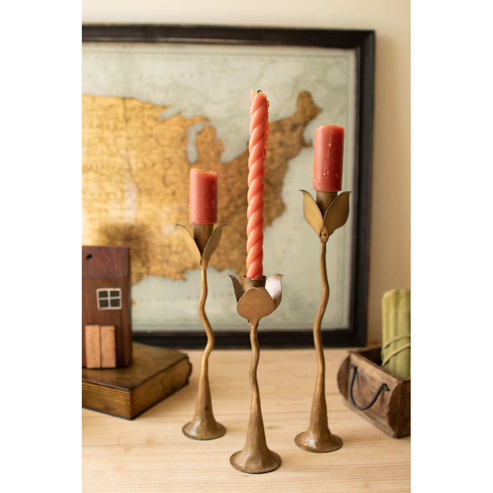 Set of three brass leaf taper candle holders styled with pink candles in front of framed map art.