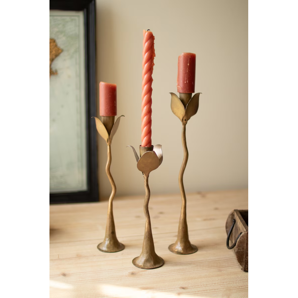Three brass leaf taper candle holders holding red candles on a light wood tabletop.
