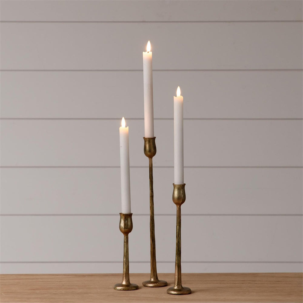 Set of 3 gold metal tapered candle holders in varying heights with lit white candles on a wooden table.